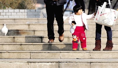 昨天中午，一女孩拉著家長的手在紫荊山公園曬太陽，氣溫升高，孩子的棉衣解開了扣子。